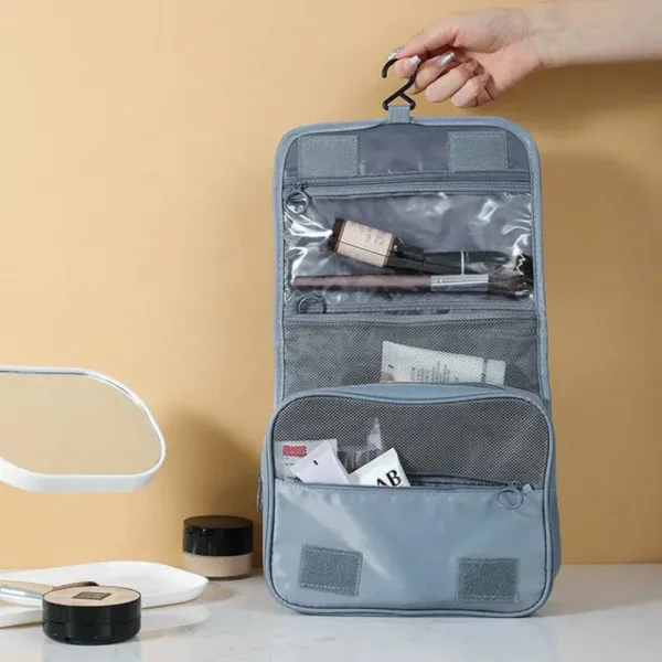 Persoană ținând de cârlig geanta de cosmetice gri deschisă complet, prezentând cele trei compartimente principale.