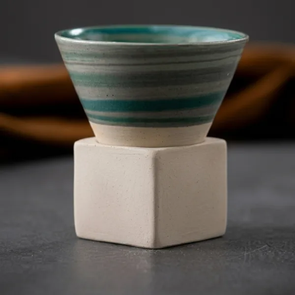 Ceasca conica din ceramica cu dungi verde smarald asezata in suportul cubic crem pe fond inchis.