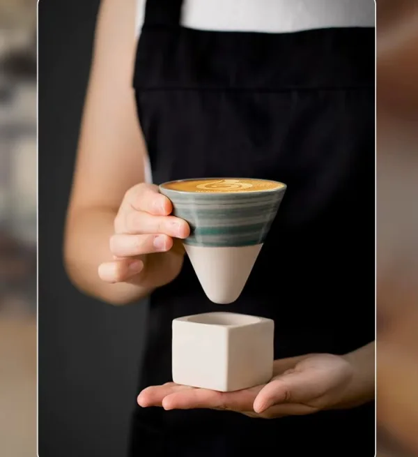 Barista tinand in mana ceasca conica verde cu latte art deasupra suportului ceramic cubic.