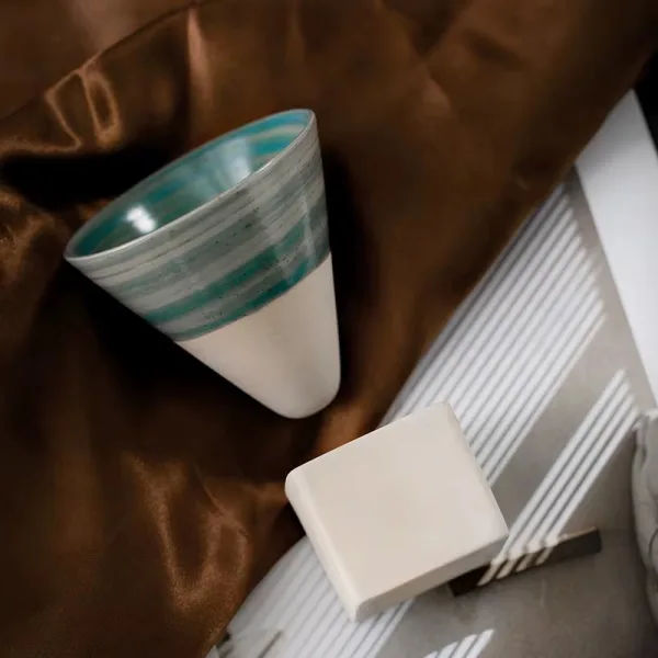 Ceasca conica de espresso cu dungi verzi si suportul sau cubic din ceramica asezate pe un material satinat maro.