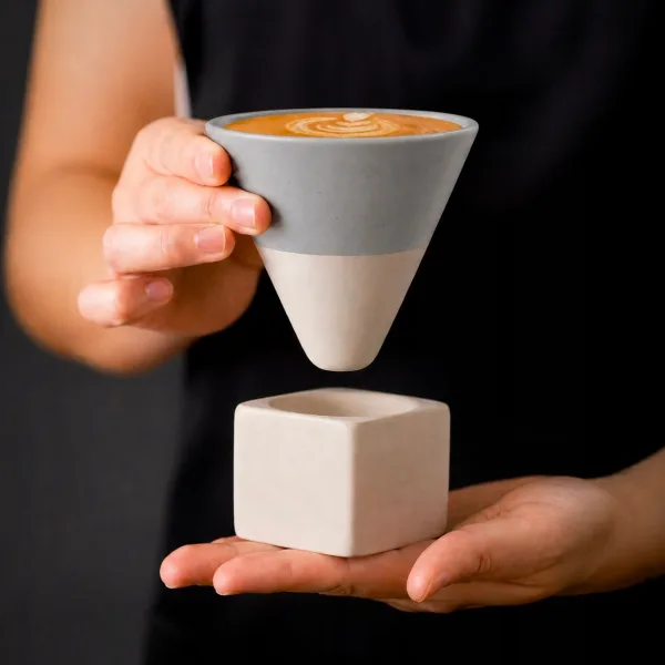 Barista tinand in mana ceasca conica gri plina cu cafea deasupra suportului ceramic cubic.