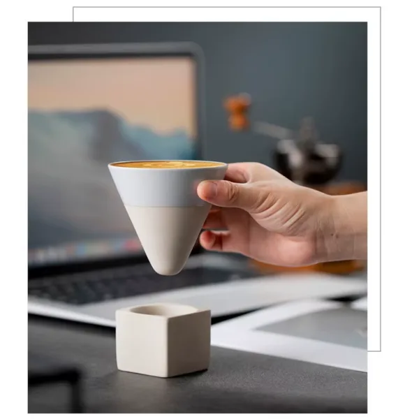 Persoana ridicand ceasca conica gri cu cafea de pe suportul cubic asezat pe un birou langa laptop.