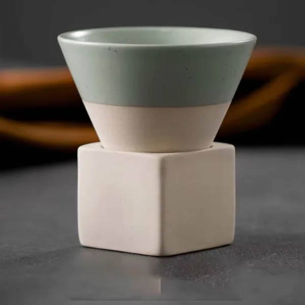 Ceasca conica din ceramica verde cu baza crem-bej asezata pe suportul cubic ceramic, pe fundal inchis.